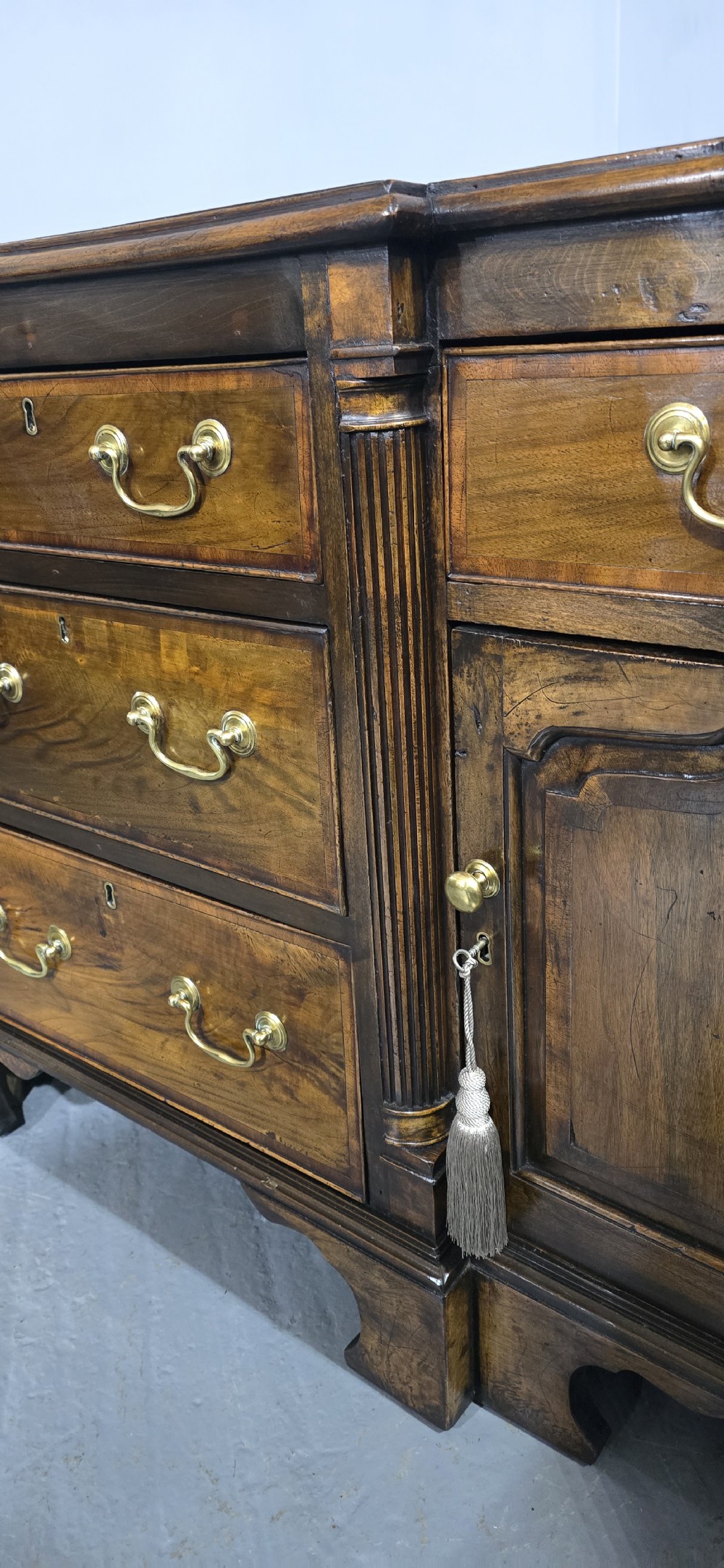 A dark oak Georgian breakfront dresser base featuring brass swan-neck handles, three central drawers, and two outer cupboard doors set on bracket feet. View VII