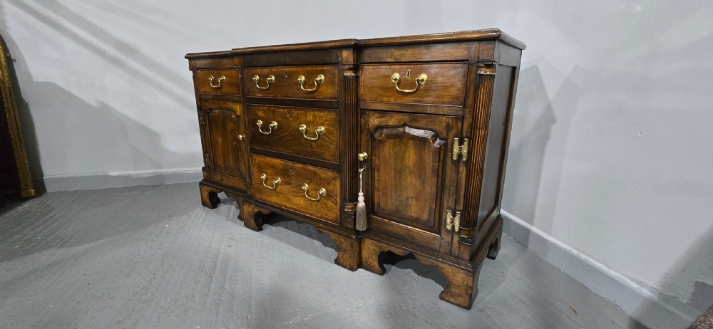 A dark oak Georgian breakfront dresser base featuring brass swan-neck handles, three central drawers, and two outer cupboard doors set on bracket feet. View XIV
