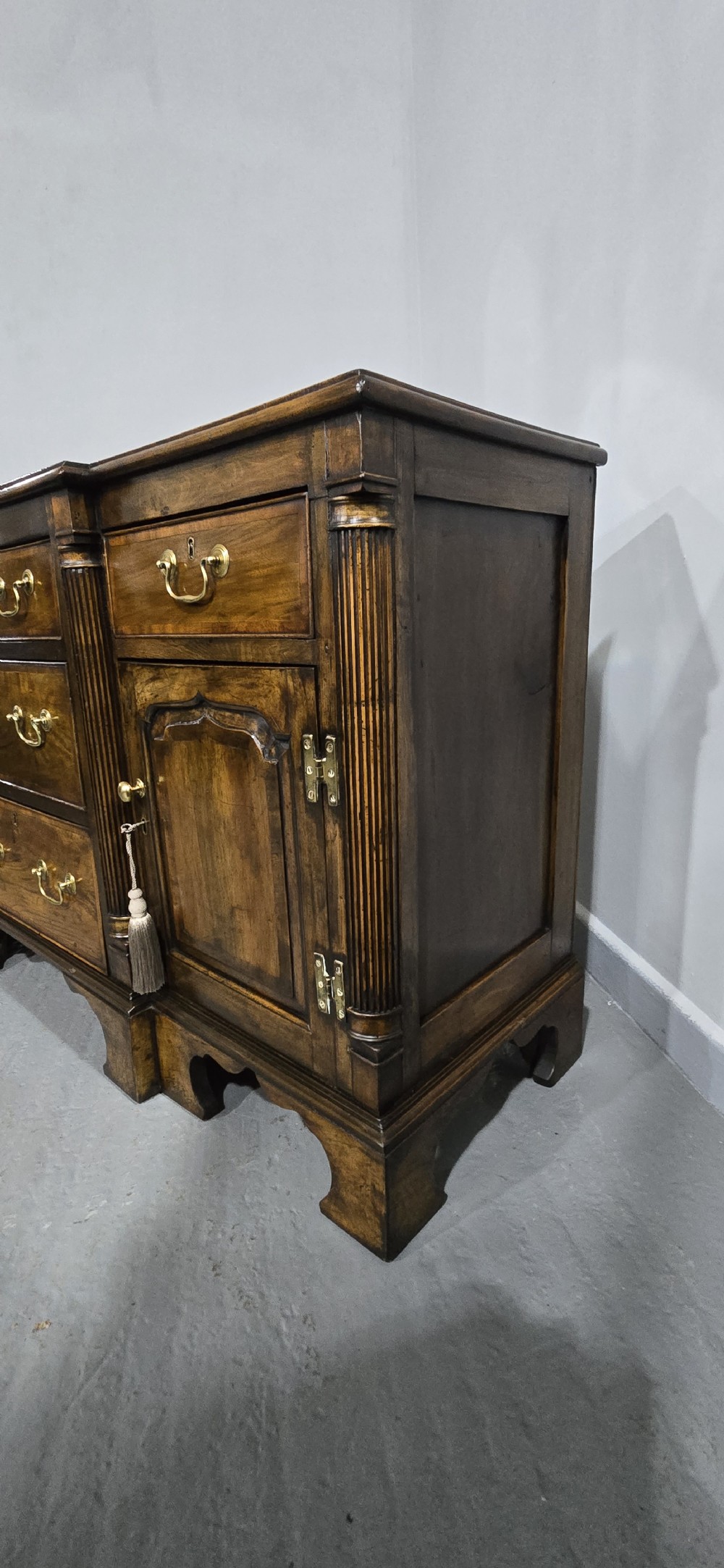 A dark oak Georgian breakfront dresser base featuring brass swan-neck handles, three central drawers, and two outer cupboard doors set on bracket feet. View XV