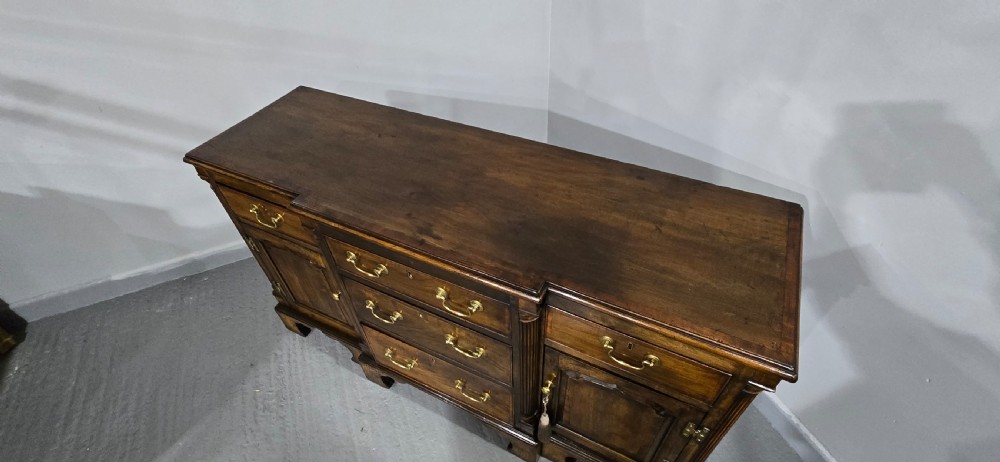 A dark oak Georgian breakfront dresser base featuring brass swan-neck handles, three central drawers, and two outer cupboard doors set on bracket feet. View XVI