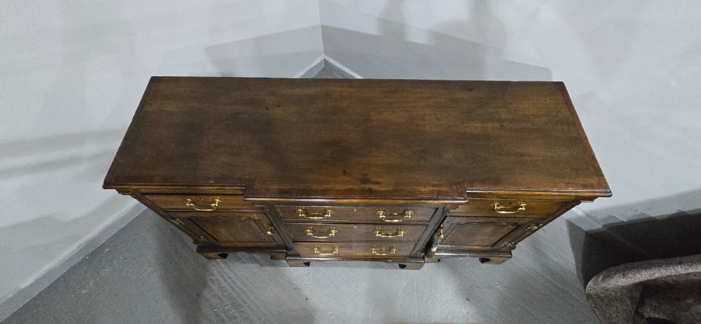 A dark oak Georgian breakfront dresser base featuring brass swan-neck handles, three central drawers, and two outer cupboard doors set on bracket feet. View XVII