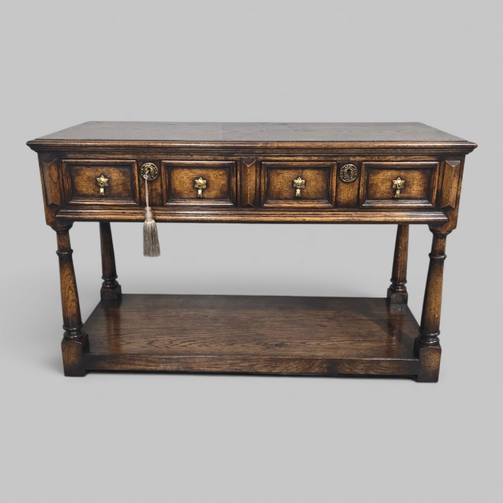 An antique dark oak dresser base featuring four panelled drawers with brass drop handles above turned supports and an open lower shelf.