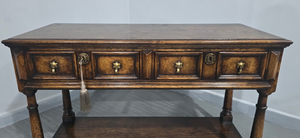 An antique dark oak dresser base featuring four panelled drawers with brass drop handles above turned supports and an open lower shelf. View III