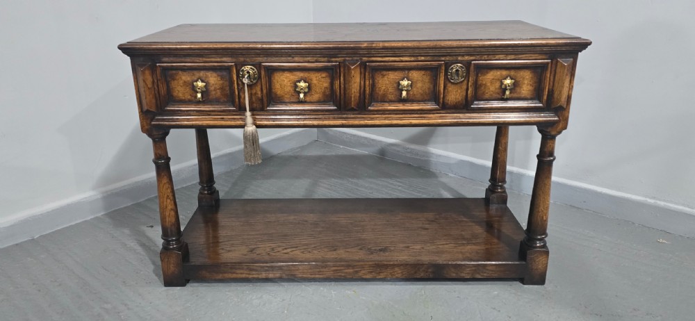 An antique dark oak dresser base featuring four panelled drawers with brass drop handles above turned supports and an open lower shelf. View IV