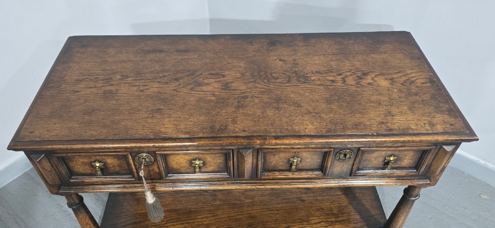 An antique dark oak dresser base featuring four panelled drawers with brass drop handles above turned supports and an open lower shelf. View V