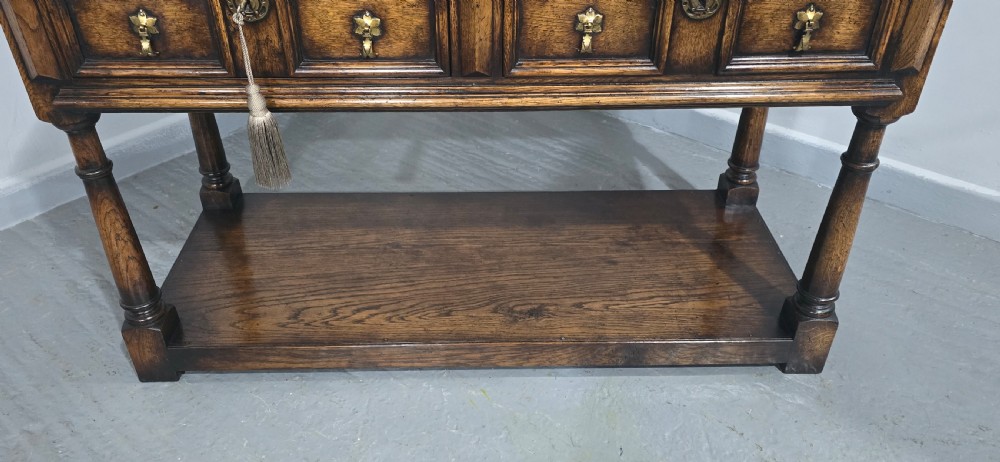 An antique dark oak dresser base featuring four panelled drawers with brass drop handles above turned supports and an open lower shelf. View VI