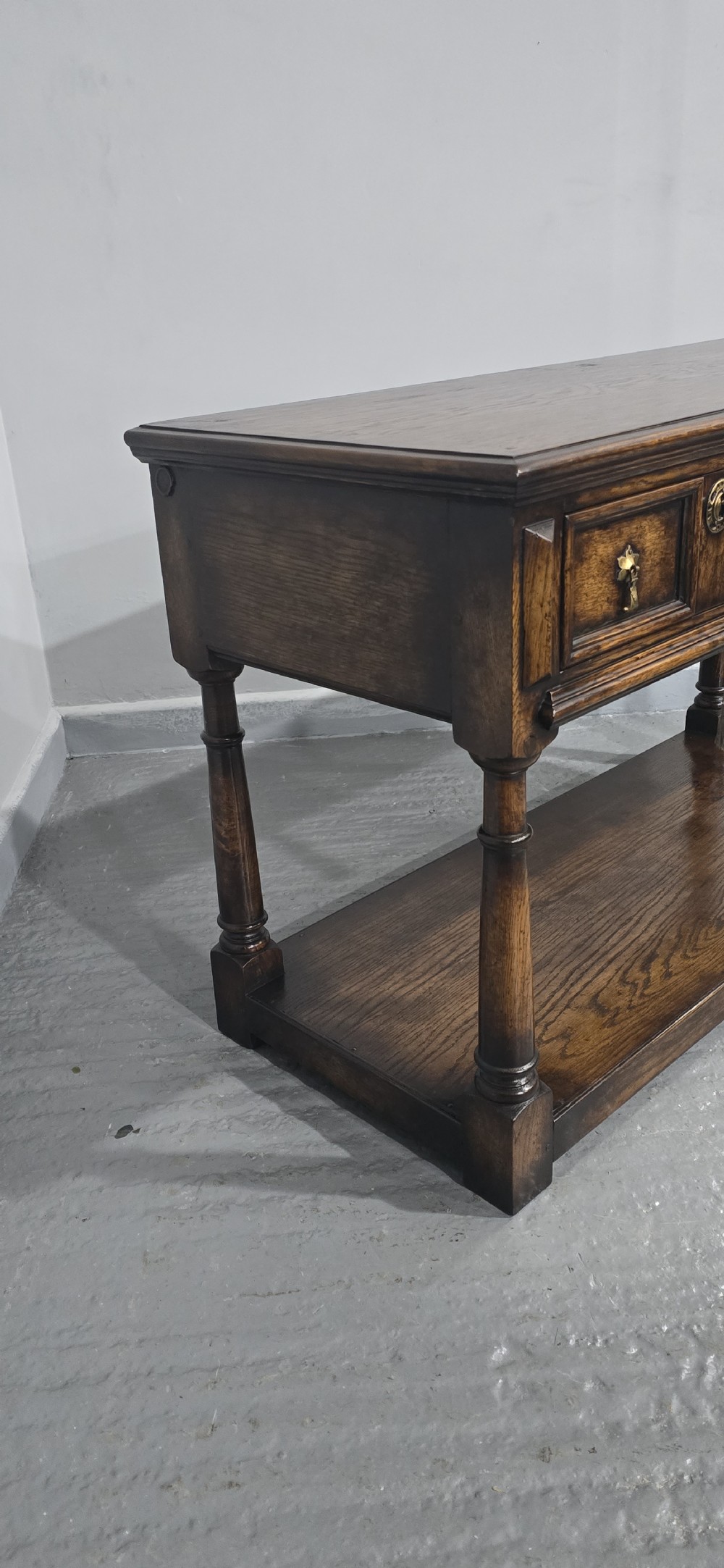 An antique dark oak dresser base featuring four panelled drawers with brass drop handles above turned supports and an open lower shelf. View IX