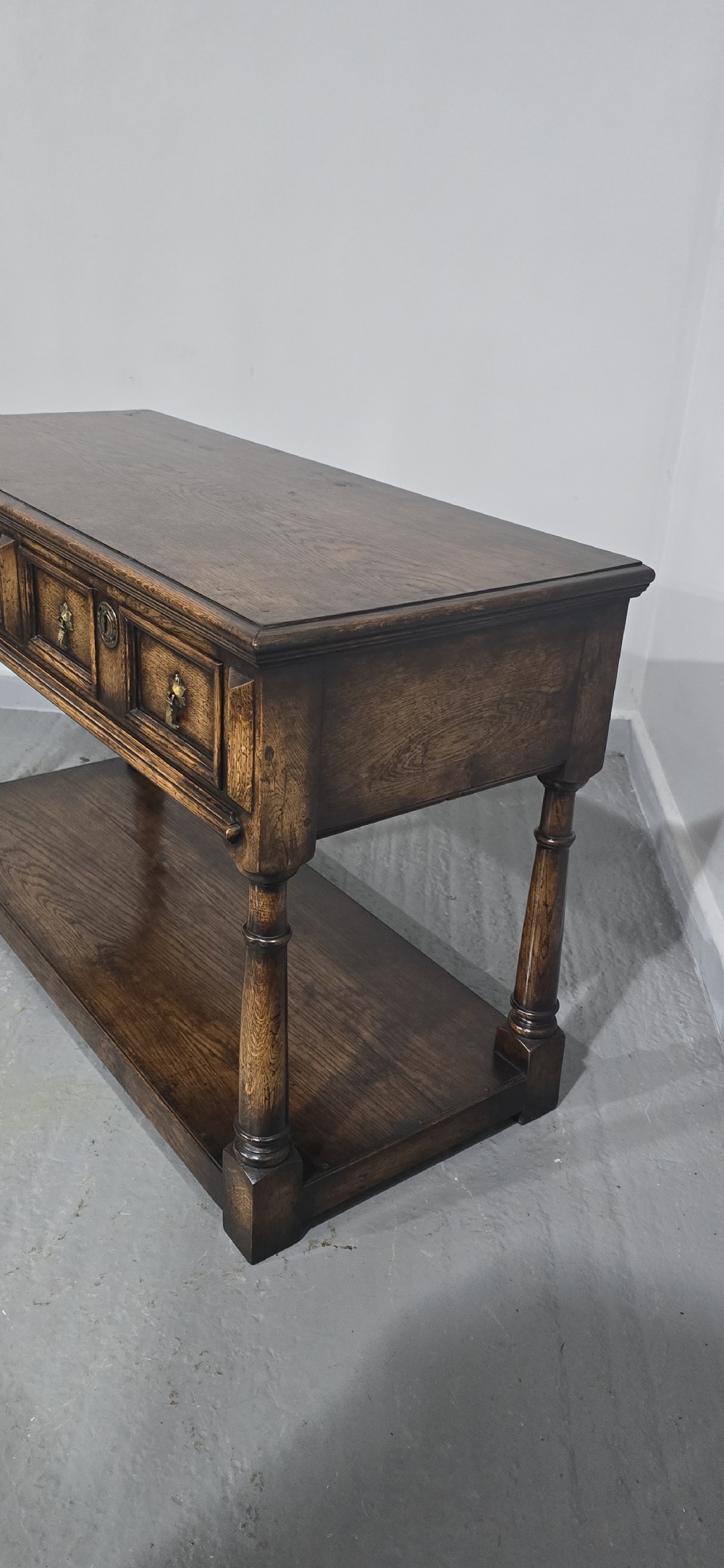 An antique dark oak dresser base featuring four panelled drawers with brass drop handles above turned supports and an open lower shelf. View X