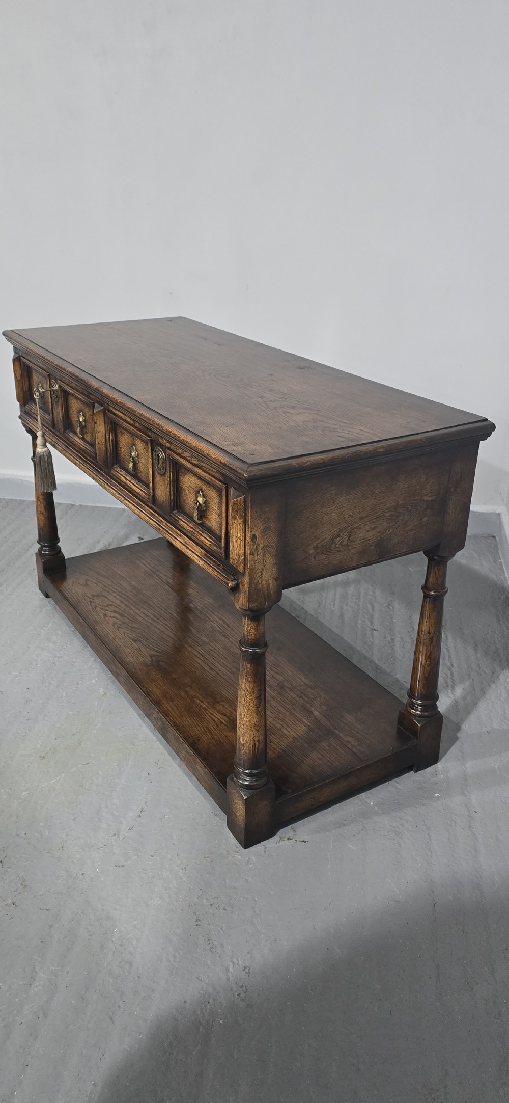 An antique dark oak dresser base featuring four panelled drawers with brass drop handles above turned supports and an open lower shelf. View XI