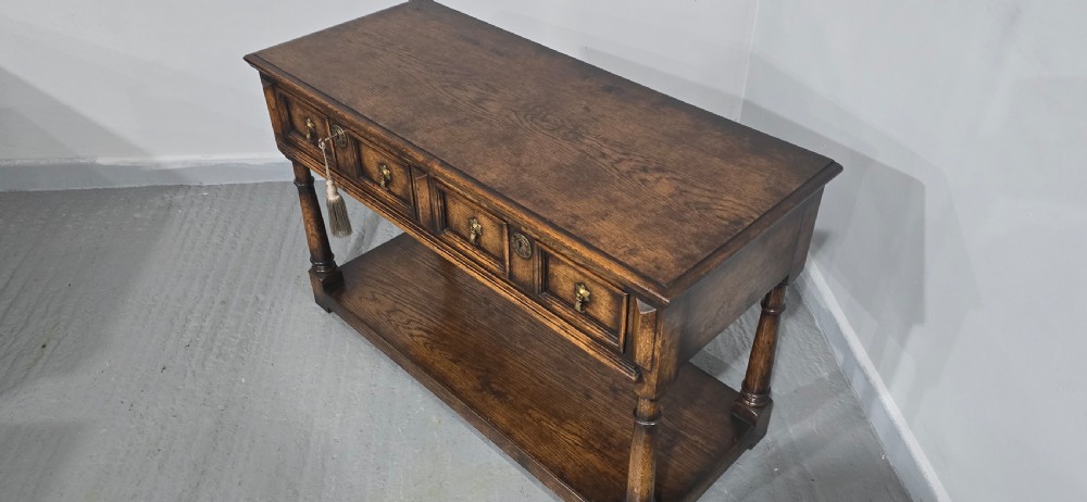 An antique dark oak dresser base featuring four panelled drawers with brass drop handles above turned supports and an open lower shelf. View XII
