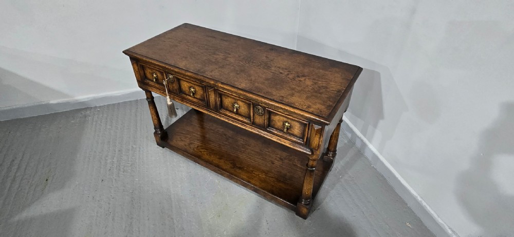 An antique dark oak dresser base featuring four panelled drawers with brass drop handles above turned supports and an open lower shelf. View XIII