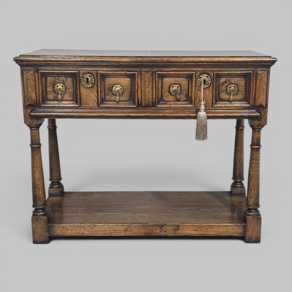 A dark oak dresser base featuring two geometric panelled drawers with brass handles, turned front legs, and a bottom shelf.