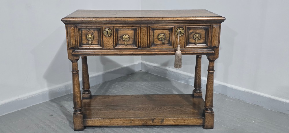A dark oak dresser base featuring two geometric panelled drawers with brass handles, turned front legs, and a bottom shelf. View III