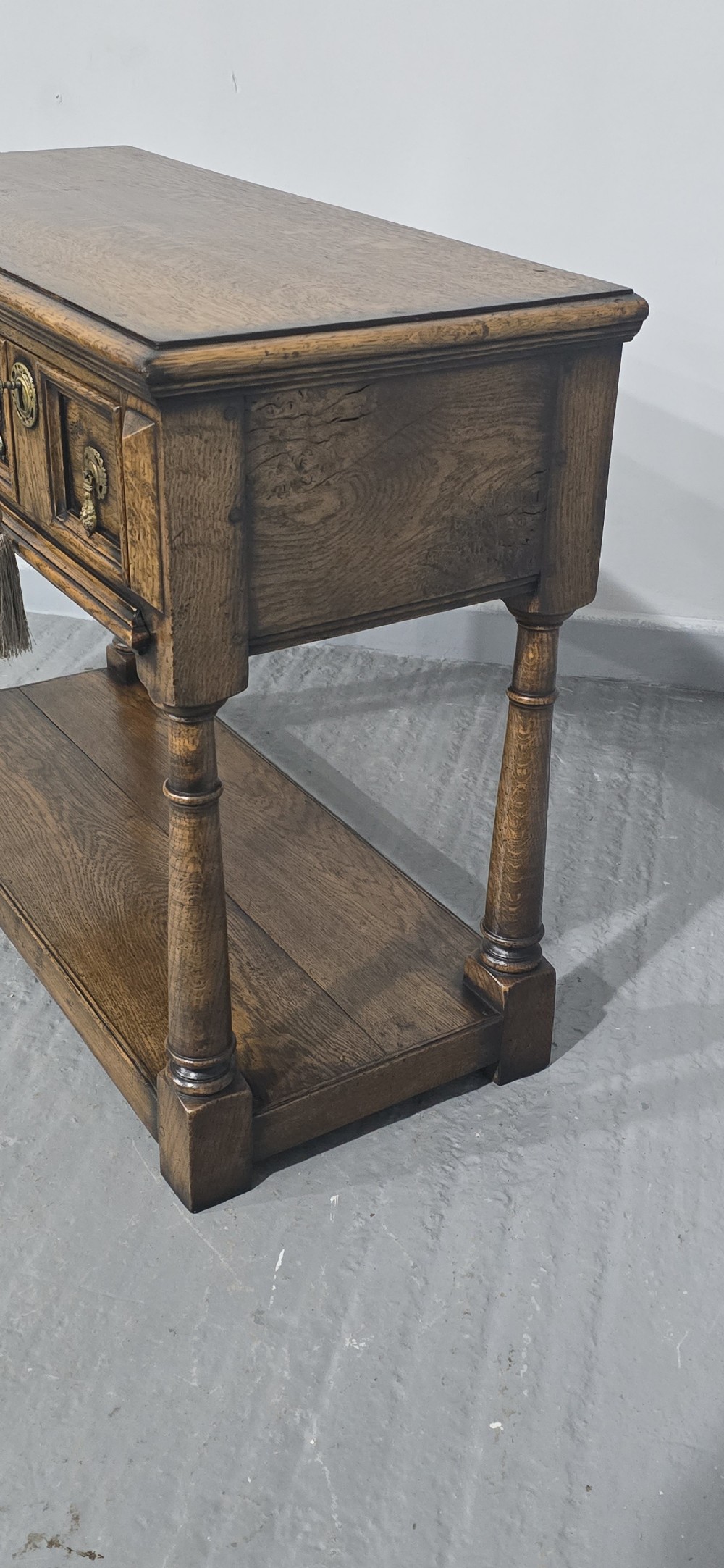 A dark oak dresser base featuring two geometric panelled drawers with brass handles, turned front legs, and a bottom shelf. View VII