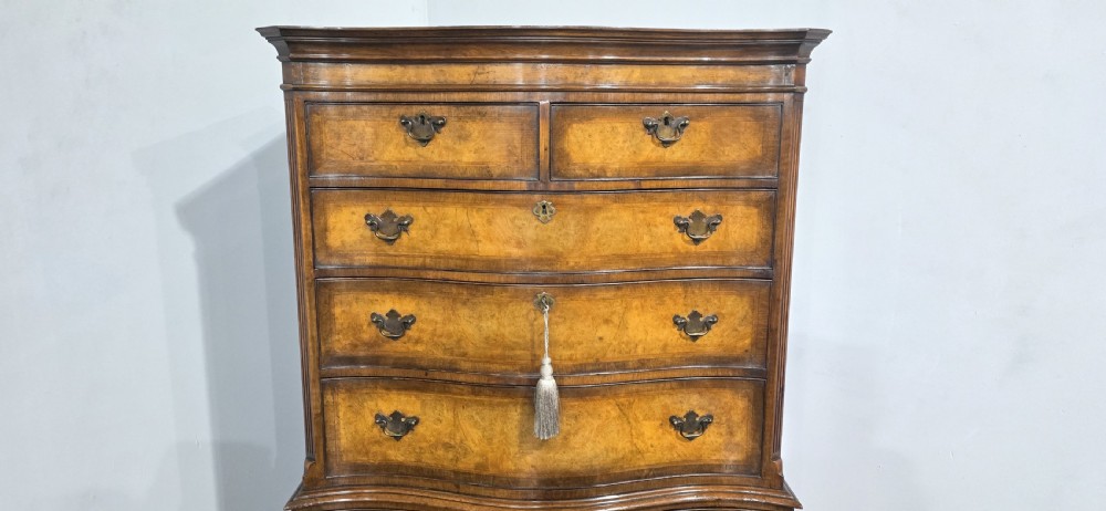 A tall two-part burr walnut chest on chest featuring a curved serpentine front, multi-drawer arrangement with brass drop handles, and bracket feet. View VI