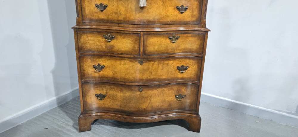 A tall two-part burr walnut chest on chest featuring a curved serpentine front, multi-drawer arrangement with brass drop handles, and bracket feet. View VII