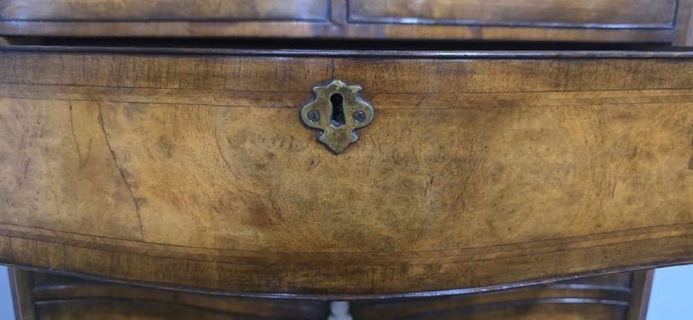 A tall two-part burr walnut chest on chest featuring a curved serpentine front, multi-drawer arrangement with brass drop handles, and bracket feet. View X