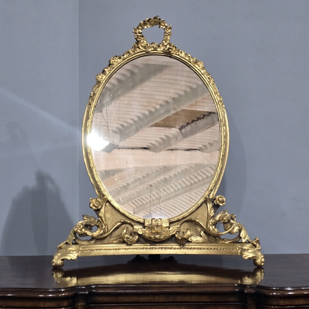 An oval gilt metal dressing table mirror with a top floral wreath cresting and an acanthus leaf scroll base. View II