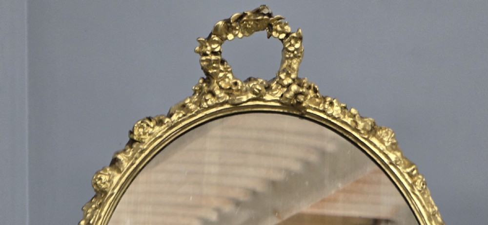 An oval gilt metal dressing table mirror with a top floral wreath cresting and an acanthus leaf scroll base. View V
