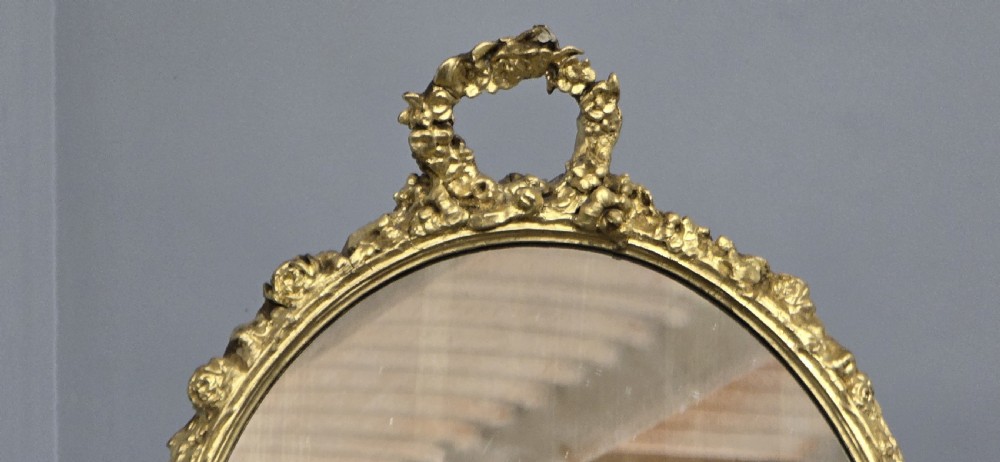 An oval gilt metal dressing table mirror with a top floral wreath cresting and an acanthus leaf scroll base. View VII
