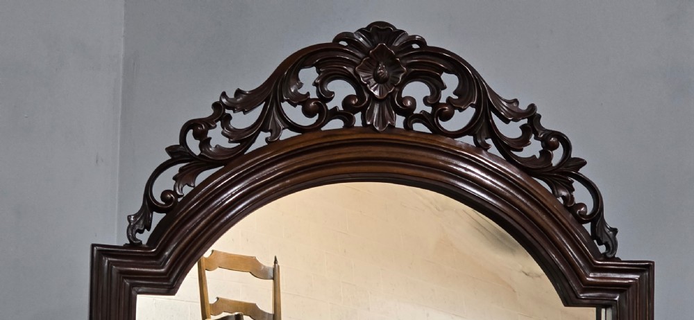 A tall mahogany cheval mirror featuring an arched frame topped with a carved scrollwork crest, mounted between two turned posts on splayed legs. View III