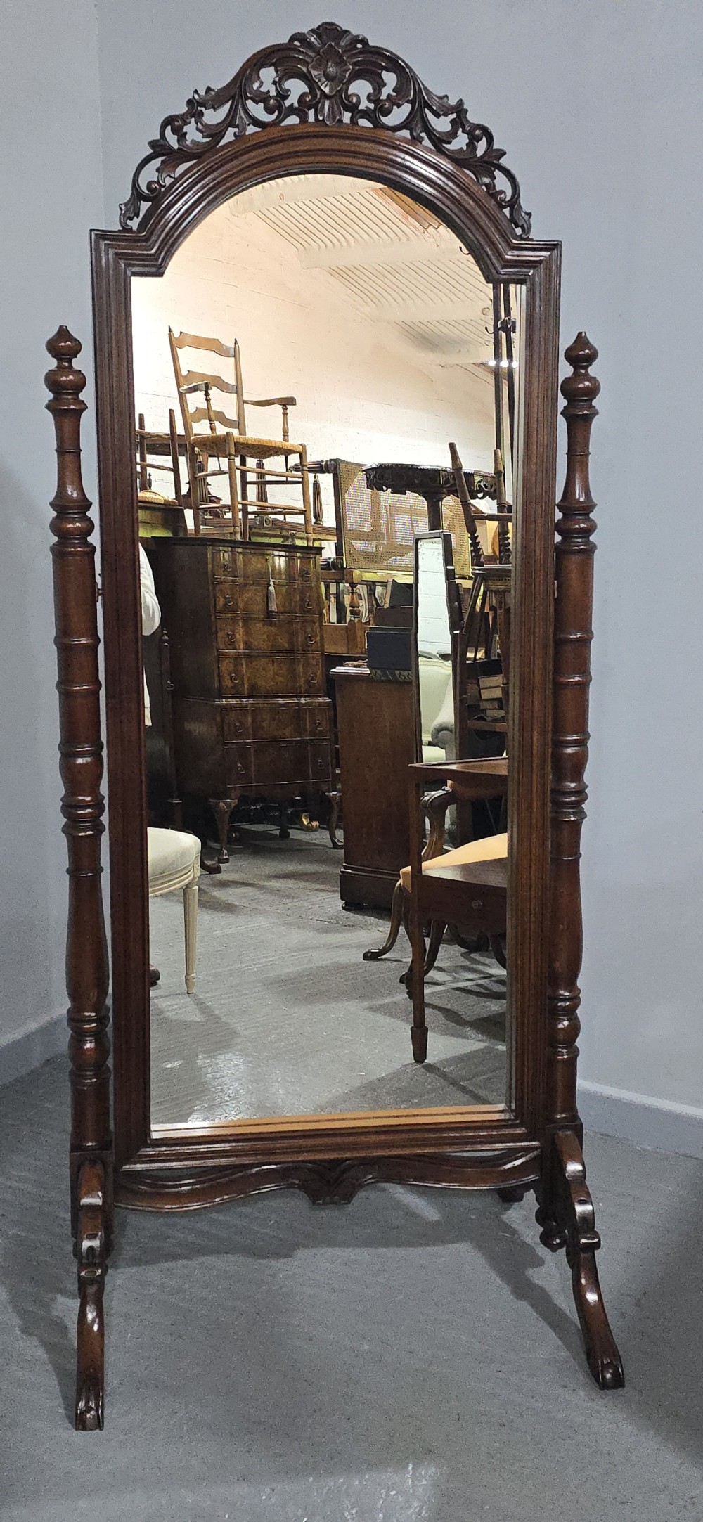 A tall mahogany cheval mirror featuring an arched frame topped with a carved scrollwork crest, mounted between two turned posts on splayed legs. View IV