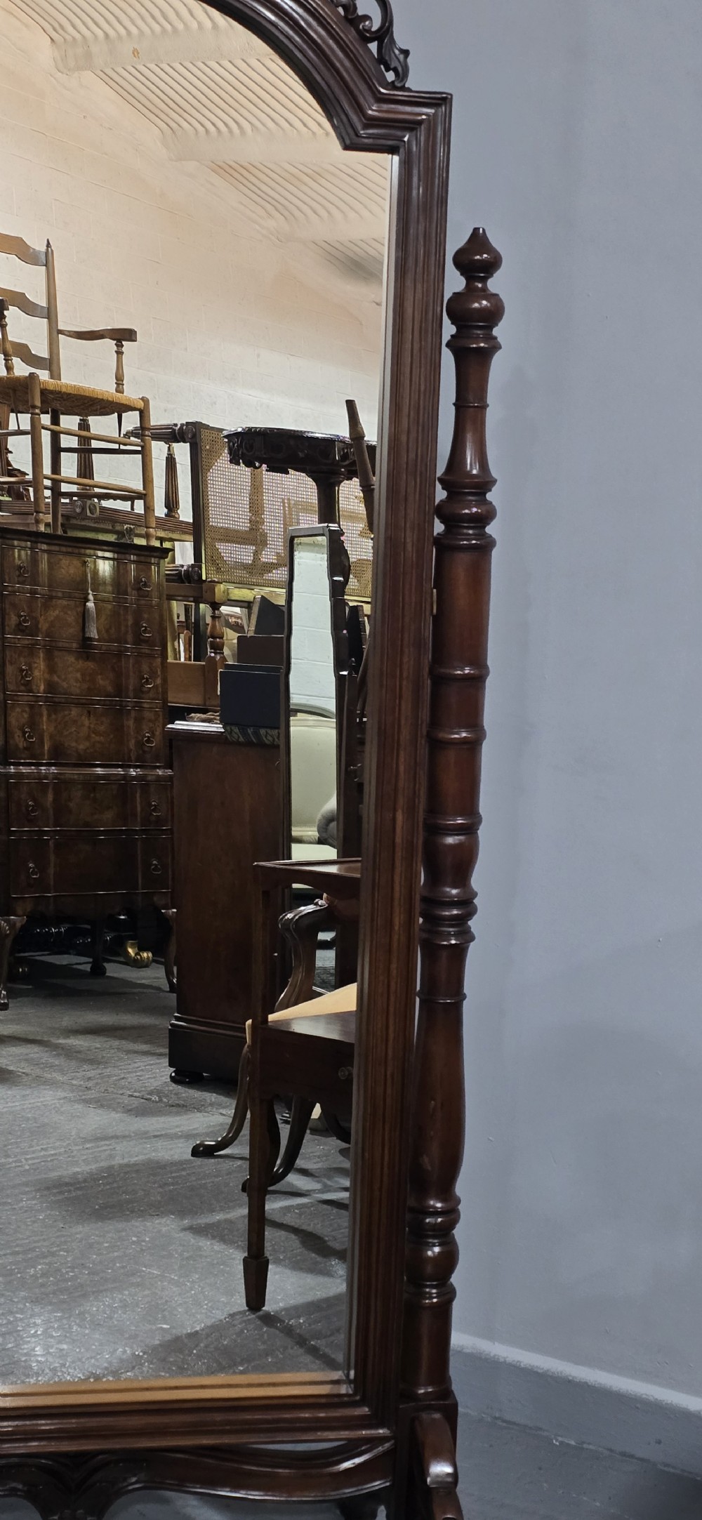 A tall mahogany cheval mirror featuring an arched frame topped with a carved scrollwork crest, mounted between two turned posts on splayed legs. View VI