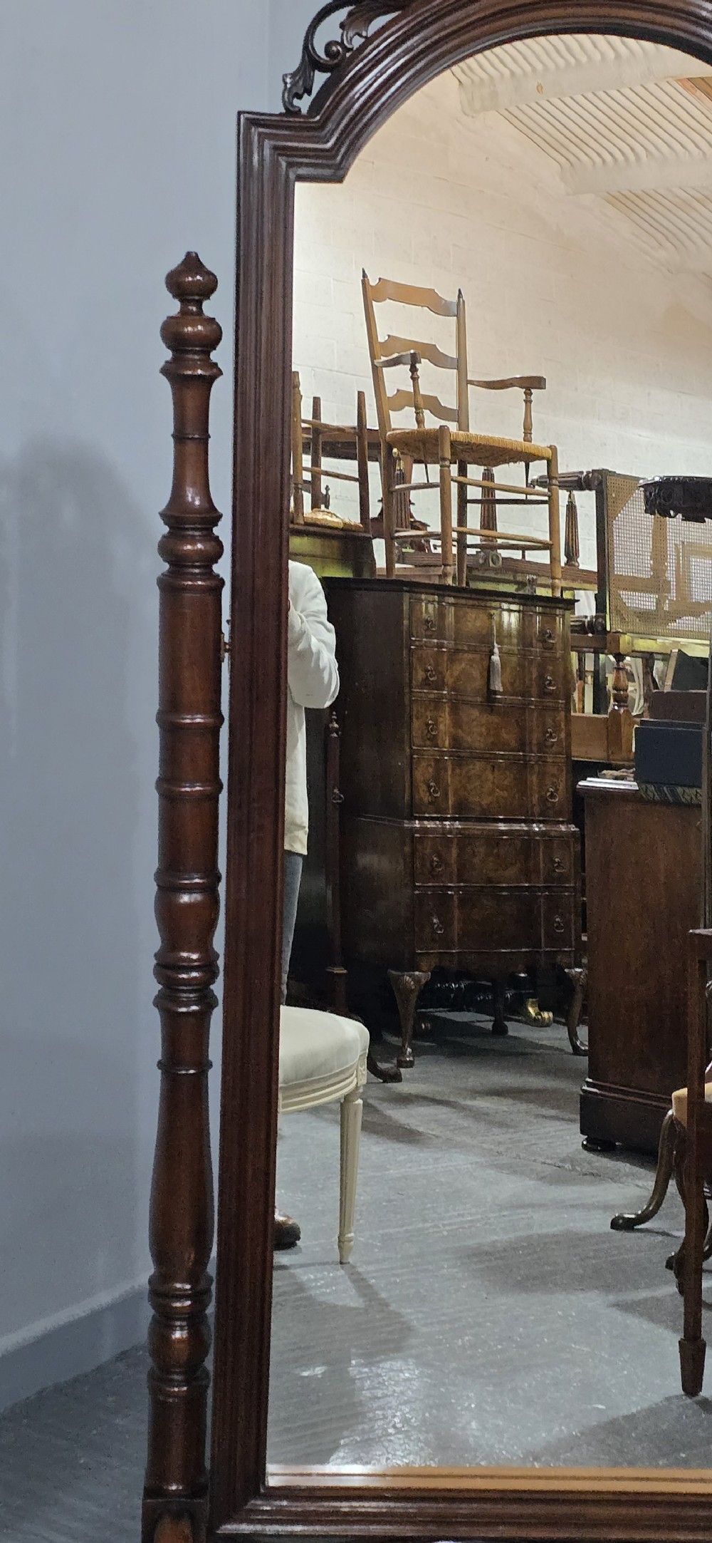 A tall mahogany cheval mirror featuring an arched frame topped with a carved scrollwork crest, mounted between two turned posts on splayed legs. View VII