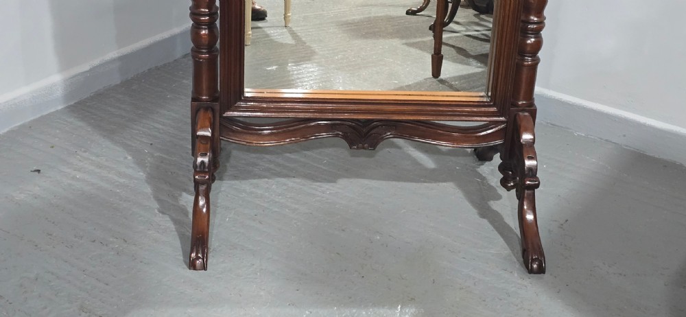 A tall mahogany cheval mirror featuring an arched frame topped with a carved scrollwork crest, mounted between two turned posts on splayed legs. View VIII