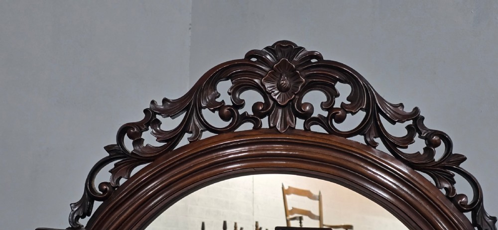 A tall mahogany cheval mirror featuring an arched frame topped with a carved scrollwork crest, mounted between two turned posts on splayed legs. View IX