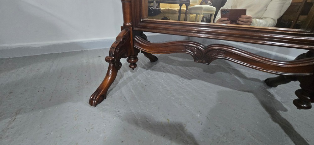 A tall mahogany cheval mirror featuring an arched frame topped with a carved scrollwork crest, mounted between two turned posts on splayed legs. View XI