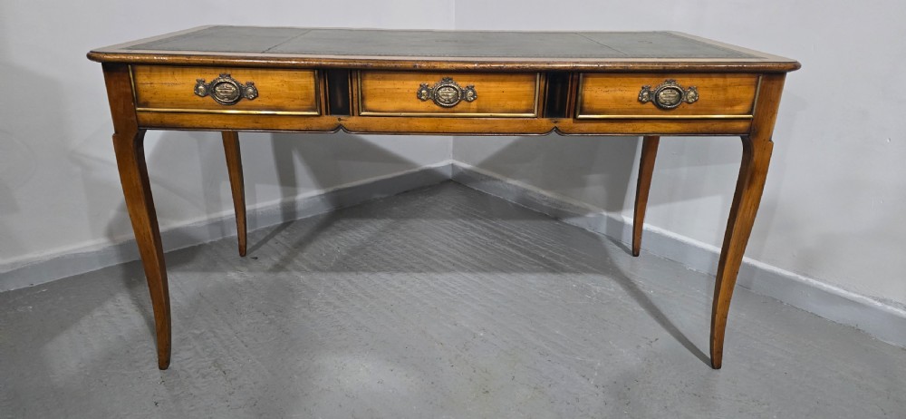 A French style warm oak library desk featuring a green leather inset writing surface, three frieze drawers with decorative brass handles, and slender cabriole legs. View X