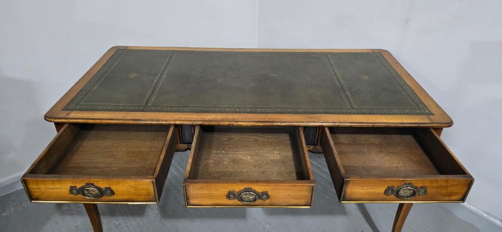 A French style warm oak library desk featuring a green leather inset writing surface, three frieze drawers with decorative brass handles, and slender cabriole legs. View XI