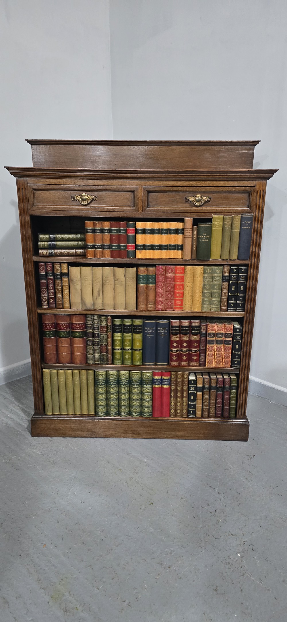 An antique oak open bookcase featuring four adjustable shelves filled with leather-bound books, twin drawers with brass handles, and fluted column side details. View III