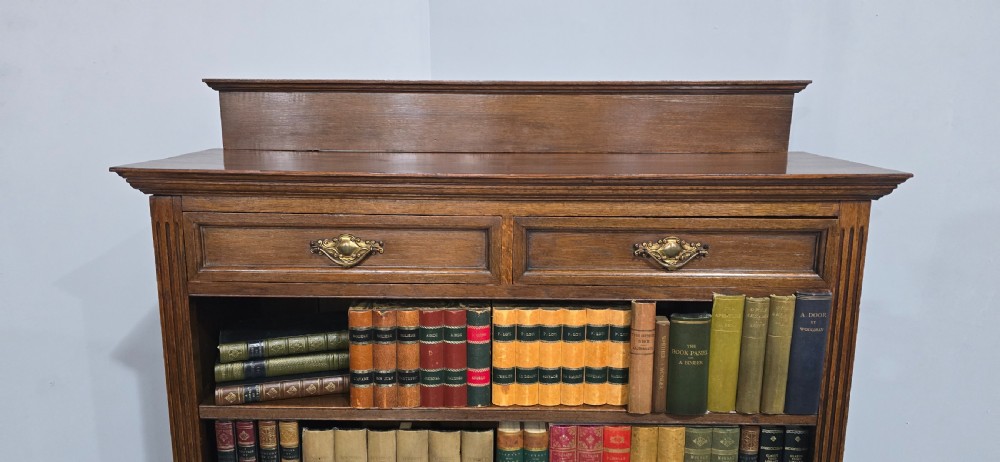An antique oak open bookcase featuring four adjustable shelves filled with leather-bound books, twin drawers with brass handles, and fluted column side details. View IV
