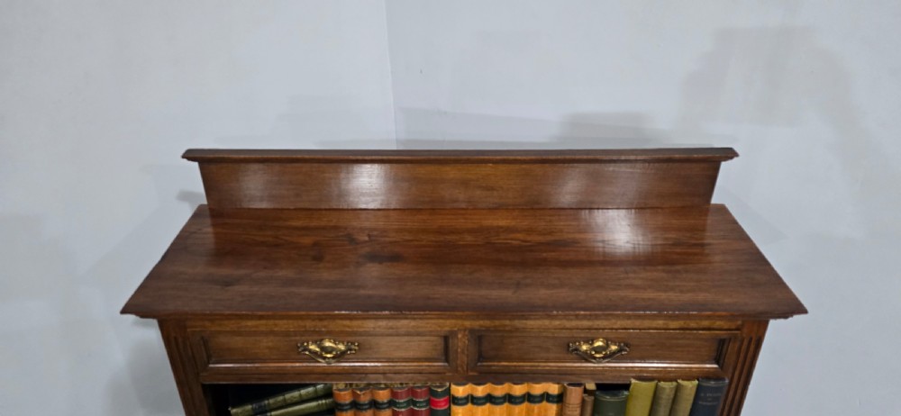 An antique oak open bookcase featuring four adjustable shelves filled with leather-bound books, twin drawers with brass handles, and fluted column side details. View V