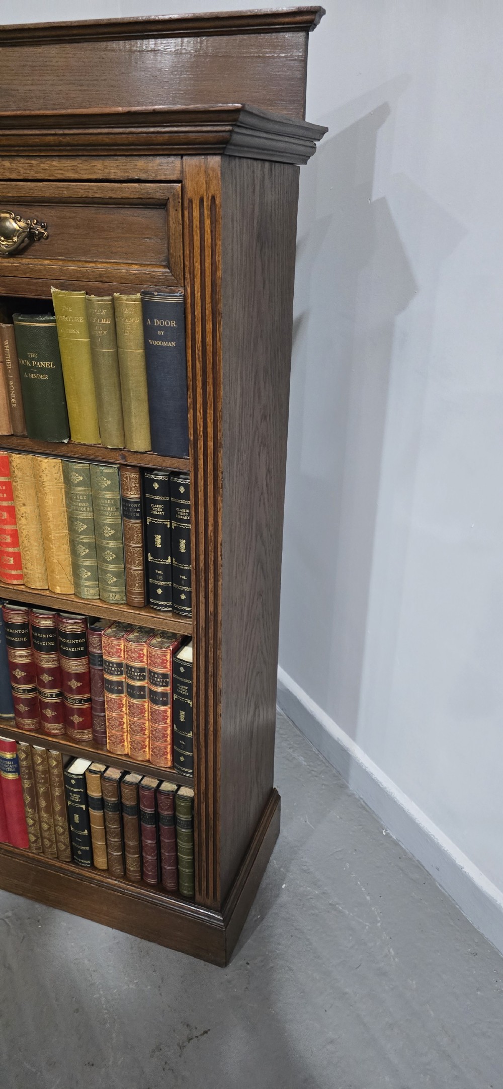 An antique oak open bookcase featuring four adjustable shelves filled with leather-bound books, twin drawers with brass handles, and fluted column side details. View VI