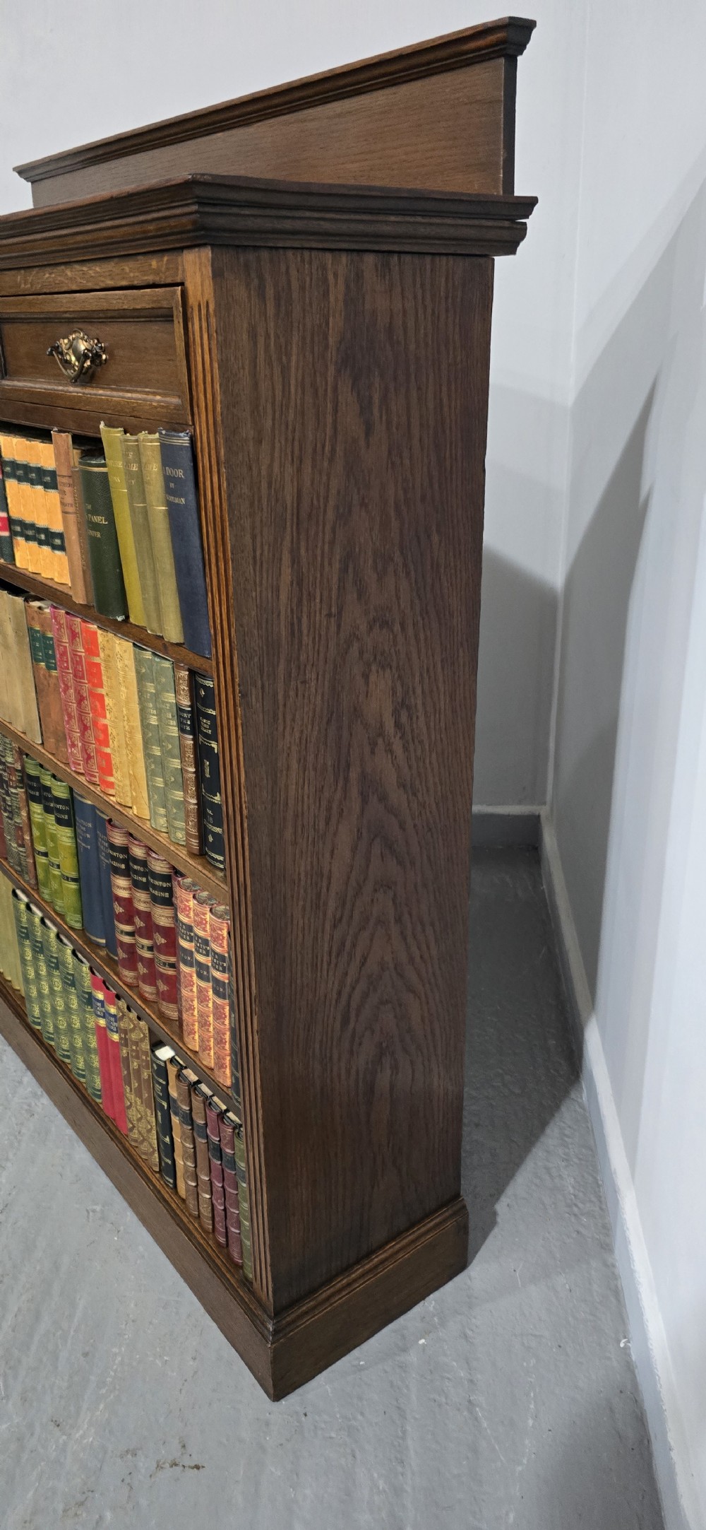 An antique oak open bookcase featuring four adjustable shelves filled with leather-bound books, twin drawers with brass handles, and fluted column side details. View VII