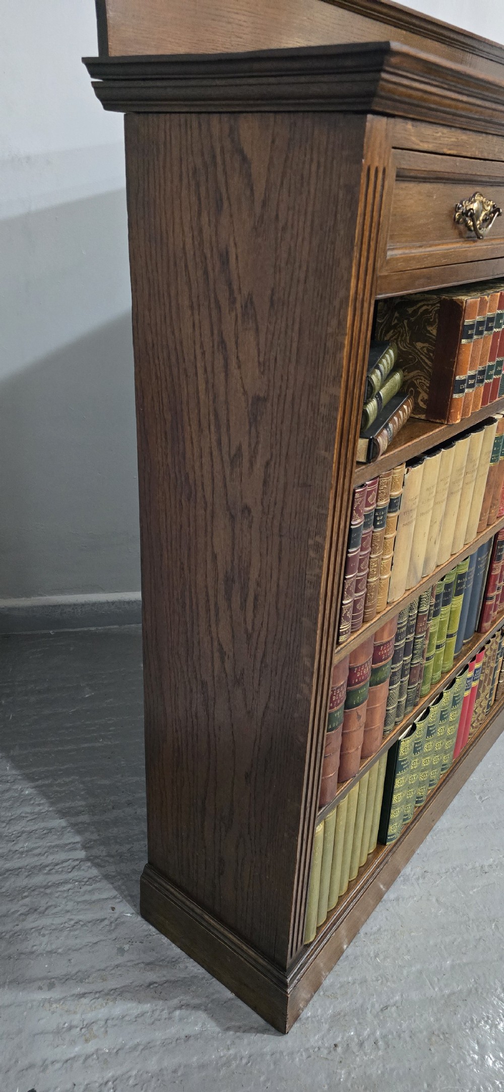 An antique oak open bookcase featuring four adjustable shelves filled with leather-bound books, twin drawers with brass handles, and fluted column side details. View VIII