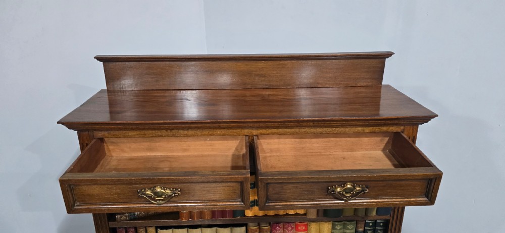 An antique oak open bookcase featuring four adjustable shelves filled with leather-bound books, twin drawers with brass handles, and fluted column side details. View X