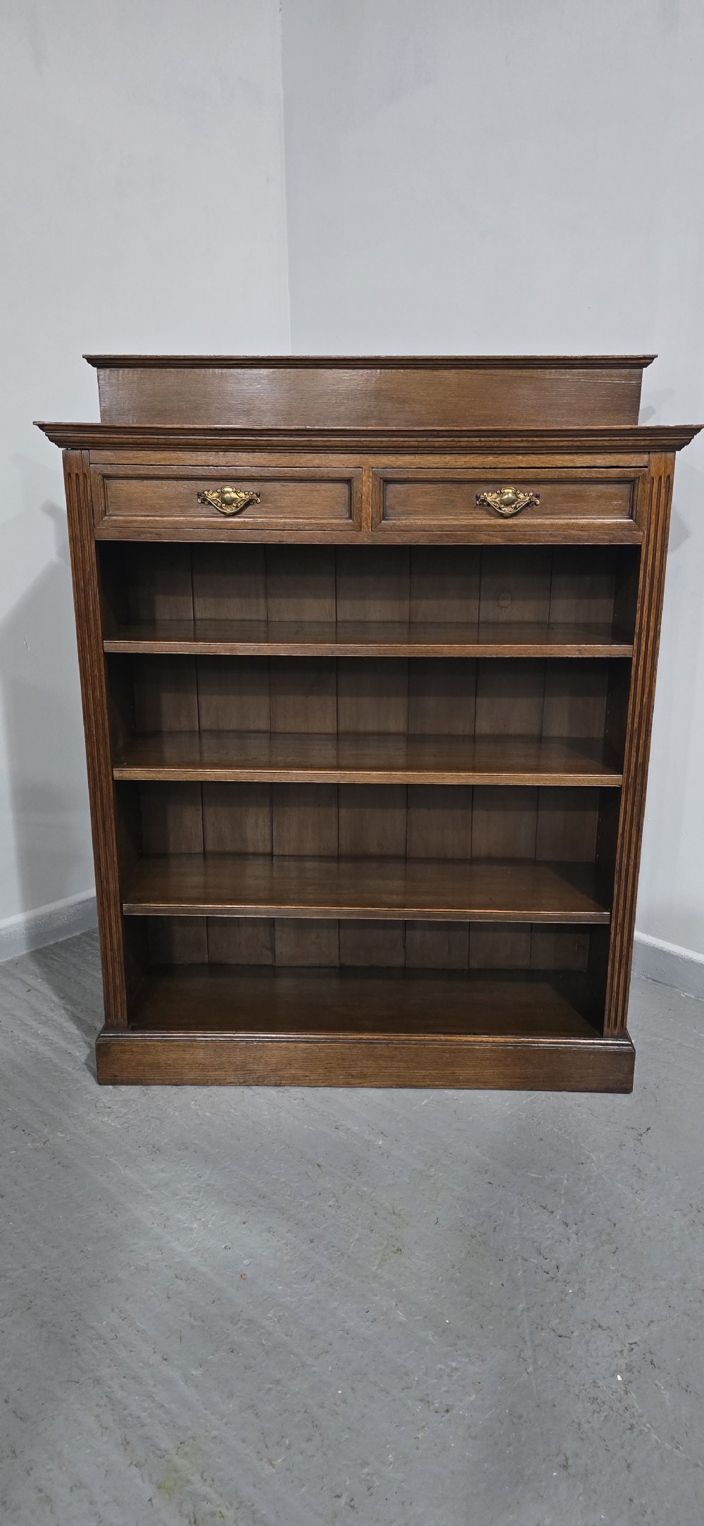 An antique oak open bookcase featuring four adjustable shelves filled with leather-bound books, twin drawers with brass handles, and fluted column side details. View XI