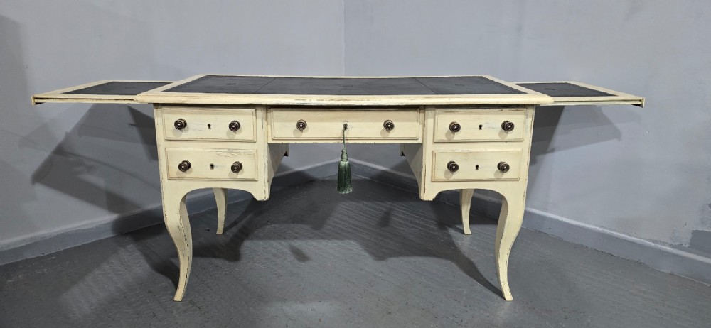 A cream painted French kneehole desk featuring a black leather inset writing surface, curved cabriole legs, and brass drawer knobs with a key and tassel hanging from the central drawer. View IV