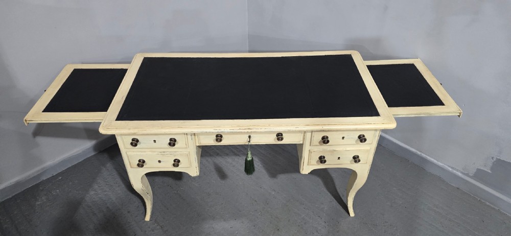 A cream painted French kneehole desk featuring a black leather inset writing surface, curved cabriole legs, and brass drawer knobs with a key and tassel hanging from the central drawer. View V