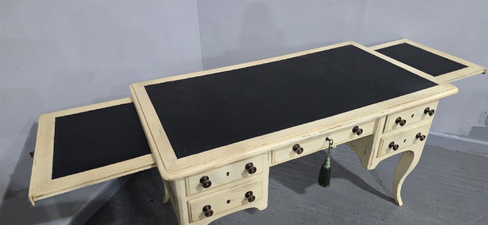A cream painted French kneehole desk featuring a black leather inset writing surface, curved cabriole legs, and brass drawer knobs with a key and tassel hanging from the central drawer. View XI