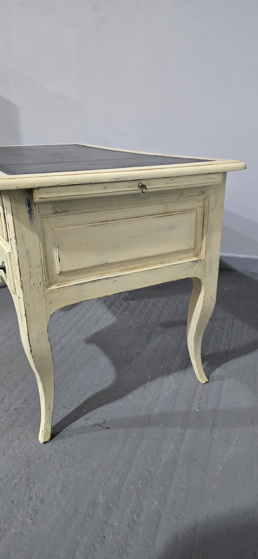 A cream painted French kneehole desk featuring a black leather inset writing surface, curved cabriole legs, and brass drawer knobs with a key and tassel hanging from the central drawer. View XV