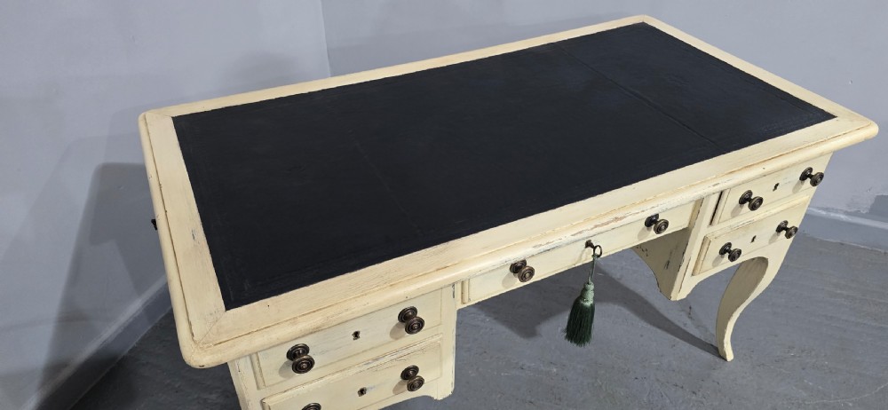 A cream painted French kneehole desk featuring a black leather inset writing surface, curved cabriole legs, and brass drawer knobs with a key and tassel hanging from the central drawer. View XVII