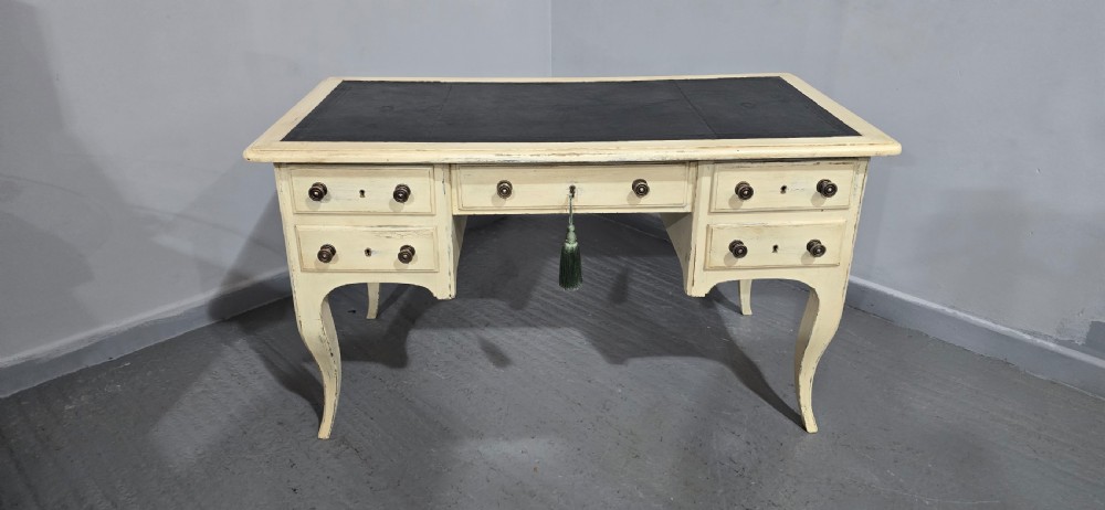 A cream painted French kneehole desk featuring a black leather inset writing surface, curved cabriole legs, and brass drawer knobs with a key and tassel hanging from the central drawer. View XVIII