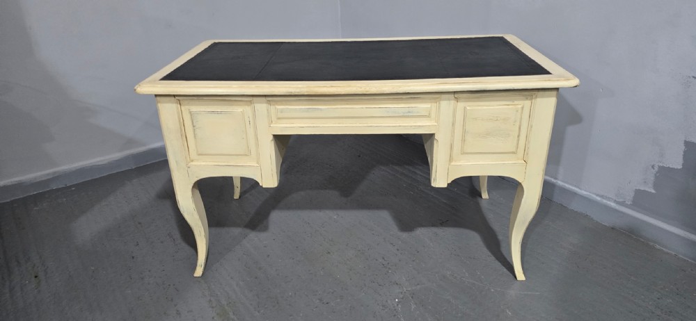 A cream painted French kneehole desk featuring a black leather inset writing surface, curved cabriole legs, and brass drawer knobs with a key and tassel hanging from the central drawer. View XIX