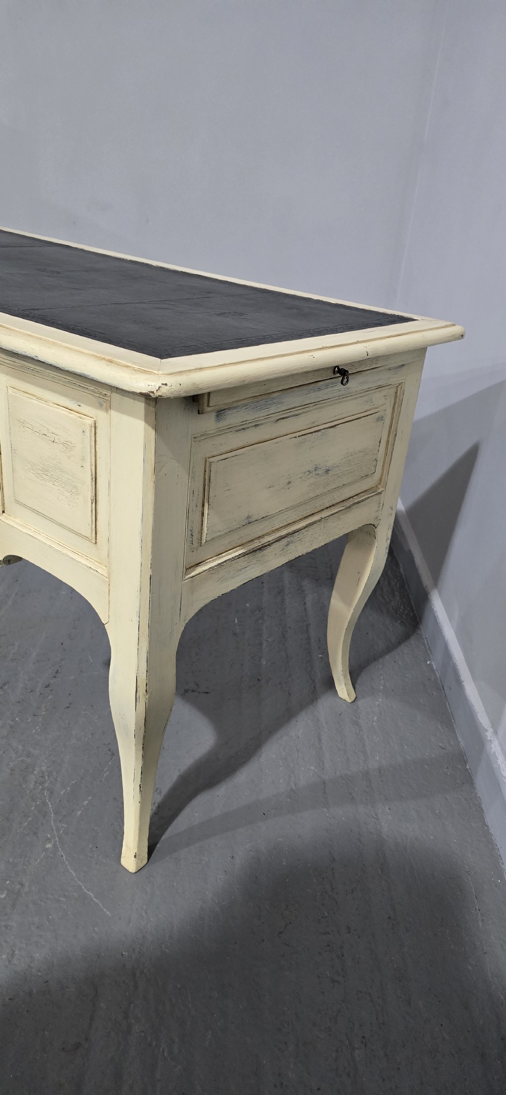 A cream painted French kneehole desk featuring a black leather inset writing surface, curved cabriole legs, and brass drawer knobs with a key and tassel hanging from the central drawer. View XX