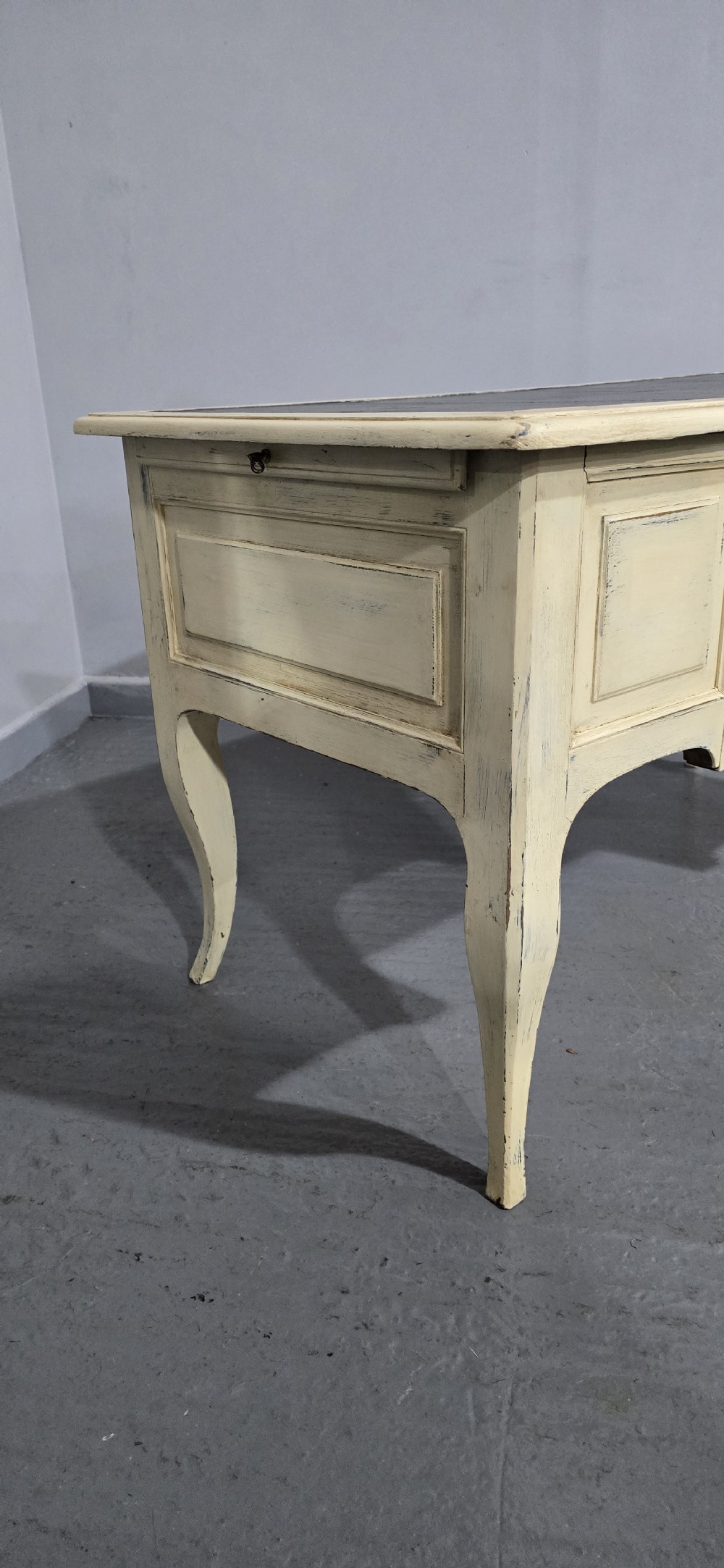 A cream painted French kneehole desk featuring a black leather inset writing surface, curved cabriole legs, and brass drawer knobs with a key and tassel hanging from the central drawer. View XXI
